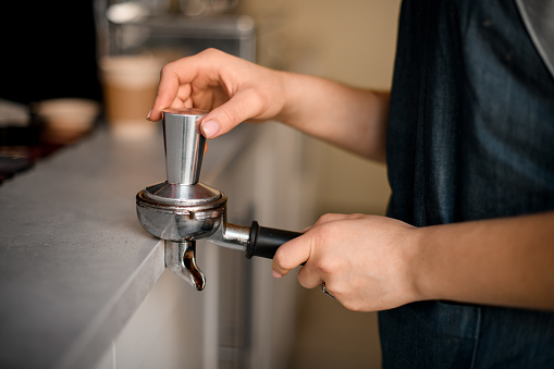 Close-up view on hand of female barista tamping coffee in portafilter before making fresh drink in coffee machine at coffee shop.