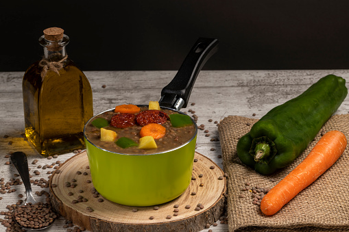 Stewed lentils with chorizo, apple or potato, green pepper and carrot in a green metal saucepan and on a wooden board. Concept of traditional and rustic cooking. selective focus