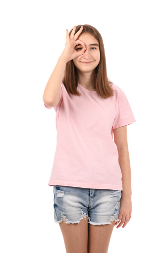 Young beautiful girl in a pink t-shirt and denim shorts on a white background, doing ok gesture with hand smiling, eye looking through fingers with happy face.