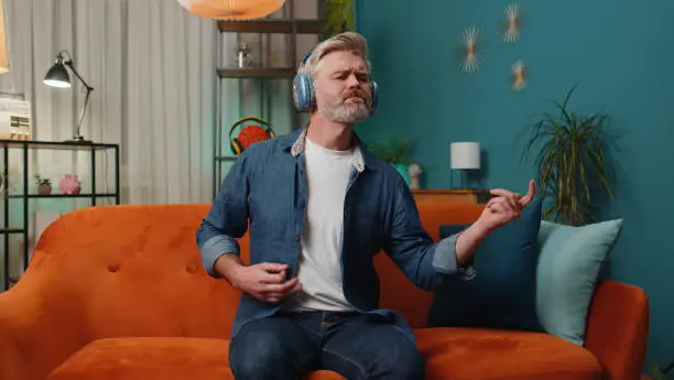 Photo of Adult man in headphones dancing on sofa at home listening favorite music playing on imaginary guitar
