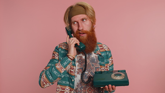Crazy hippie redhead bearded man in pattern shirt talking on wired vintage retro telephone of 80s, fooling making silly faces, unpleasant conversation. Hipster ginger guy boy on pink studio background