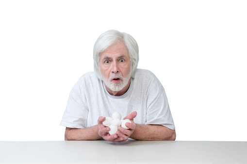 Horizontal shot of an old man sitting at a table with his hands full of eggs looking very surprised or frightened.  Isolated on white.  Lots of copy space.