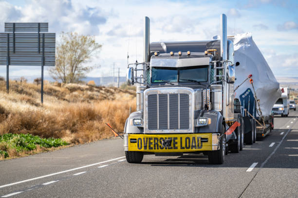Classic American big rig black semi truck with oversize load sign transporting heavy duty covered oversized cargo on step down semi trailer driving on the highway road in front of another traffic stock photo