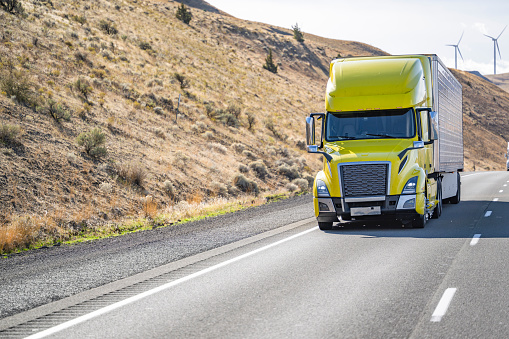 Industrial long hauler big rig yellow comfortable semi truck tractor transporting cargo in refrigerator semi trailer driving uphill on the divided highway road surrounded by hills in Washington