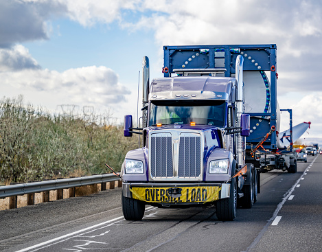 Industrial blue big rig semi truck tractor with additional trolley transports oversize load a super long blade of a wind turbine with an escort car driving on the highway road in Columbia Gorge