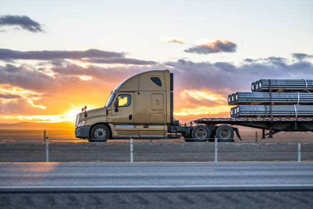 Profile of big rig beige semi truck transporting cargo on flat bed semi trailer driving on the one way highway road at sunset time stock photo
