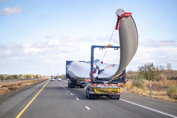 Powerful big rig semi truck with additional axle dolly with oversize load sign transporting wind turbine blade running on the straight highway road Industrial powerful big rig semi truck tractor with additional trolley transports oversize load a super long blade of a wind turbine with an escort car driving on the highway road in Columbia Gorge large machine stock pictures, royalty-free photos & images