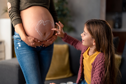 Little girl being playful drawing heart shape with body cream on her mom's belly in living room at home. Mother and daughter spending time together, enjoying moments of pure happiness.