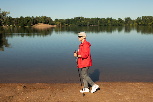Grey-haired woman walking with tracking sticks on the beach near lake, lifestyle concept.