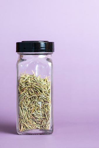 Dried rosemary leaves in a rectangular glass spice jar placed off center on a lilac colored background with copy space.