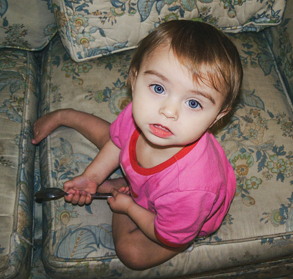 Photo has been adjusted to appear older than it is.  Toddler girl in pink top sits on beige couch with blue floral pattern.  She is looking up at the camera and holding a spoon.