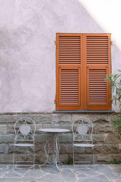 Exterior of building with stony decoration and closed window with wooden shutters near white table and chairs in Liguria
