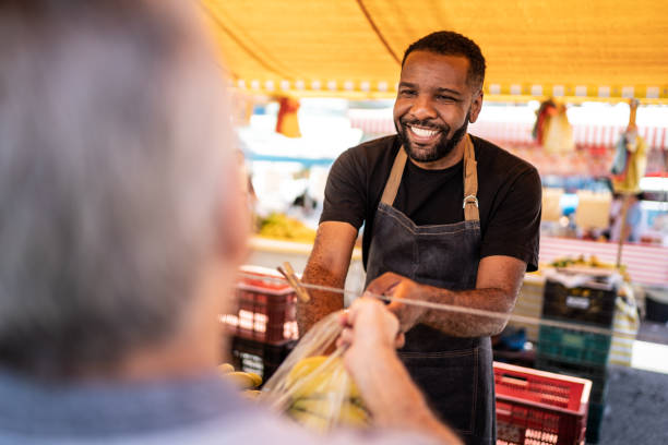 salesman helping the his customer putting the bananas in a plastic bag on a street market - kopen fotos stockfoto's en -beelden