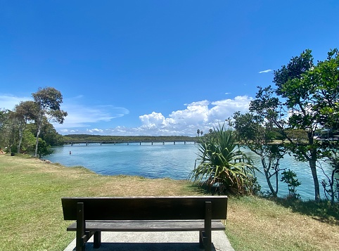 Horizontal landscape of public park bench in grass area overlooking river with coastal trees and distant foot bridge under clear blue sky at Brunswick Heads river near Byron Bay NSW Australia