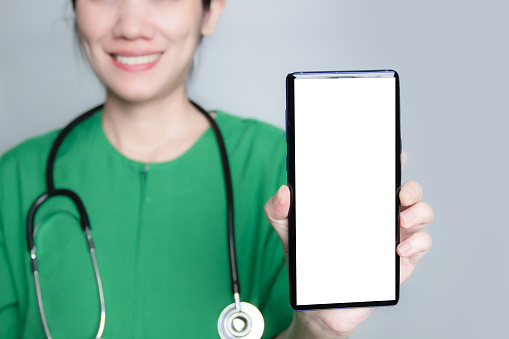 Smile and happy nurse wear green suit with stethoscope on shoulder hold and show smartphone with white screen introduce health care via a digital application.