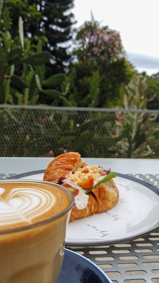A cup of hot cappuccino and tuna croissant as breakfast with green trees as the background
