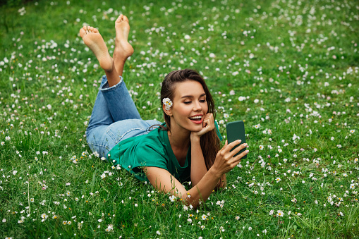 Smiling young woman lying on grass and using smartphone