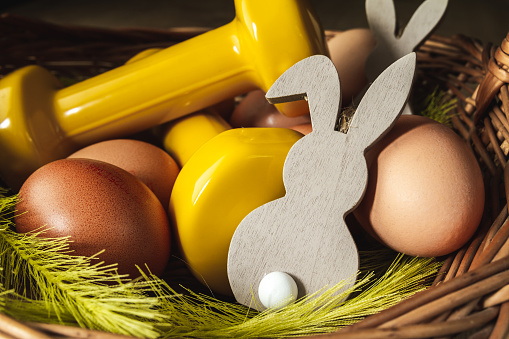 Traditional Easter wicker basket with eggs, decorative bunny and yellow dumbbells. Healthy fitness lifestyle composition, gym workout and training concept. Cheat day temptation vs sticking to diet.