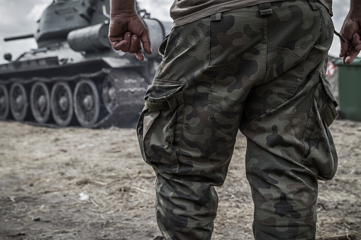 Close-up of army soldier's legs on a battlefield, wearing woodland camo military pants (camouflage trousers). Soldier with a battle tank in background.