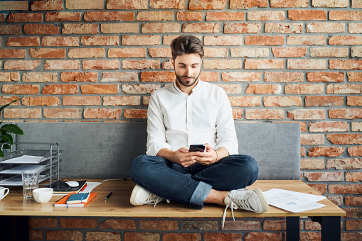 Young businessman at modern loft office sitting on desk with smartphone texting