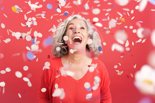 Studio portrait with red background of a cheerful mature woman surrounded by confetti in the air