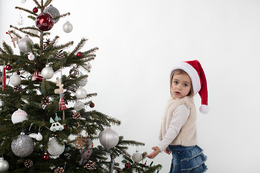 3-years old cute girl wearing Santa's hat posing next to a Christmas Tree. White background high key image