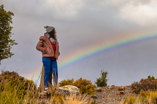 Young woman looking at rainbow from field, wearing casual clothes with hat.