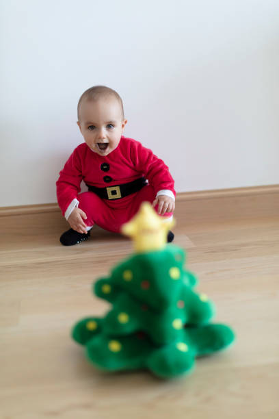 Baby girl wearing Santa's dress behind small Christmas tree soft toy. First Christmas