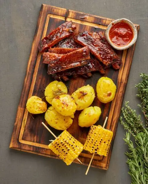 Spicy barbecue pork ribs, corn ears and crushed smashed potatoes. Slow cooking recipe. Pickled roasted pork meat with red sauce. American Cuisine.