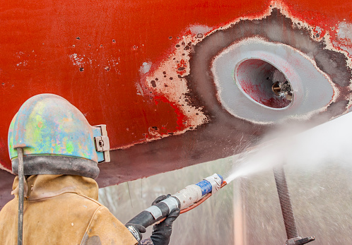 worker, sandblasting the corroded hull of a sailing vessel with a high pressure sandblasting syst
