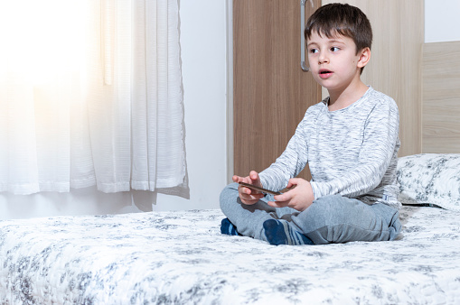 Little 6 years old boy using and playing on smartphone. Cute little boy playing games on his smartphone in bed.  Social and technology concept.