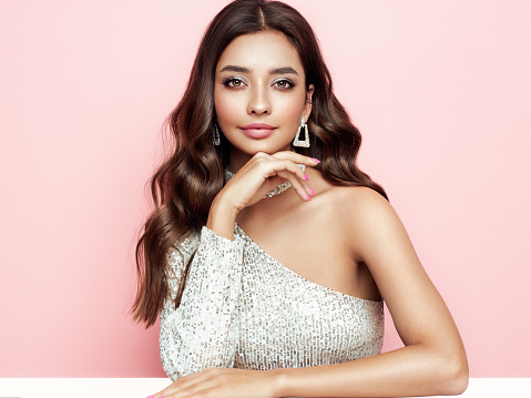 Beautiful woman in cocktail dress posing on pink background. Beauty model with long curly hair. Christmas or New Year festivities. Jewelry and manicure