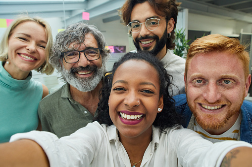 Close up portrait of young and mature employees of a multiracial team having fun and posing for a selfie with cell phone together in the office. Colleagues looking camera with cheerful expression.