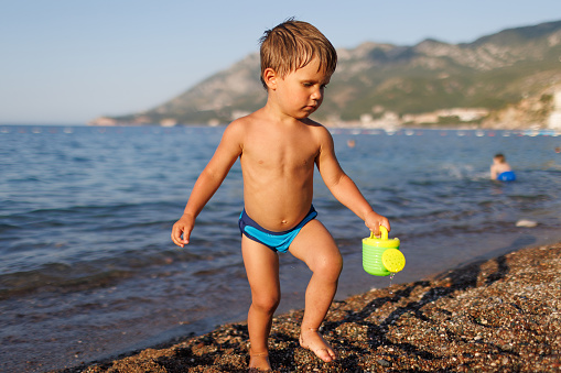 A cheerful tanned toddler boy in blue swimming trunks runs along a sandy wet beach with small shells with a bright yellow toy watering can, against the backdrop of the Adriatic Sea in the sunlight