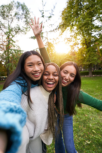 Vertical Portrait of three girls outside having fun taking selfie. An Asian Chinese woman, a black African American and a Caucasian together and embracing. Friendship in multi-ethnic groups of people