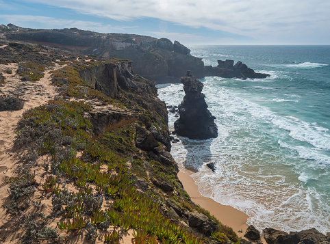 View of sharp cliffs and small sand beach with ocean waves, rocks, stones and green vegetation at wild Rota Vicentina coast near Vila Nova de Milfontes, Portugal