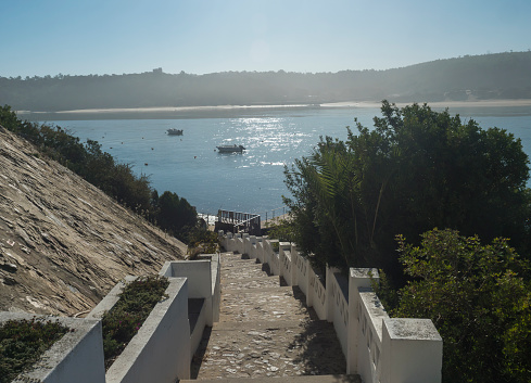 View of stairs leading to pier with small ferry boat across the river Mira flowing into the Atlantic Ocean. Vila Nova de Milfontes, Vicentine Coast Natural Park Portugal.