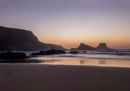 After sunset blue hour dreamy long exposure view of sand beach Praia da Zambujeira do Mar with rock and cliff and blurred ocean waves in pink light. Rota Vicentina coast, Odemira, Portugal