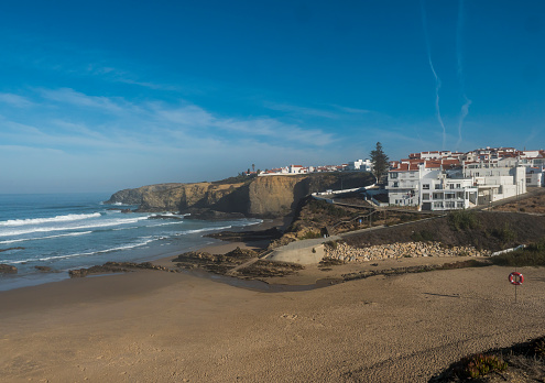 Vista de la playa de arena dorada y el pueblo de Praia da Zambujeira do Mar con olas del mar, acantilados y piedras, salvavidas rojo y vegetación verde en la costa salvaje de Rota Vicentina, Odemira, Portugal. photo