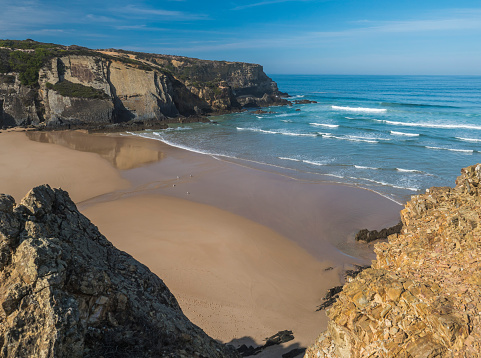 View of empty Praia do Carvalhal beach with ocean waves, cliffs and stones, wet golden sand and green vegetation at wild Rota Vicentina coast, Odemira, Portugal