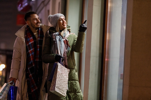 Portrait of happy young couple walking embraced down the city street, carrying shopping bags, looking inside the store window while shopping during Christmas holidays. Young woman is pointing at something inside.