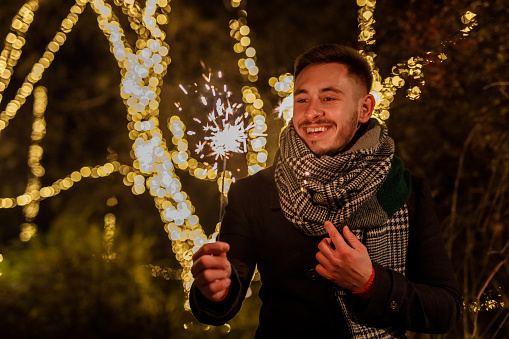 Copy space shot of cheerful young man standing at the park, against glistening Christmas lights, holding a sparkler, smiling and making a wish.