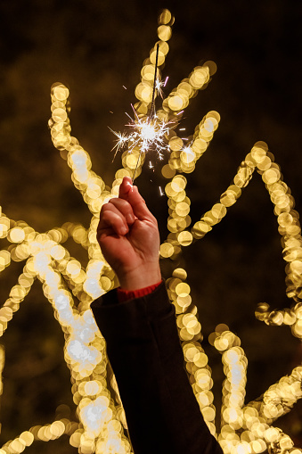 Close up shot of unrecognizable young man standing at the park, against glistening Christmas lights, waving a sparkler.