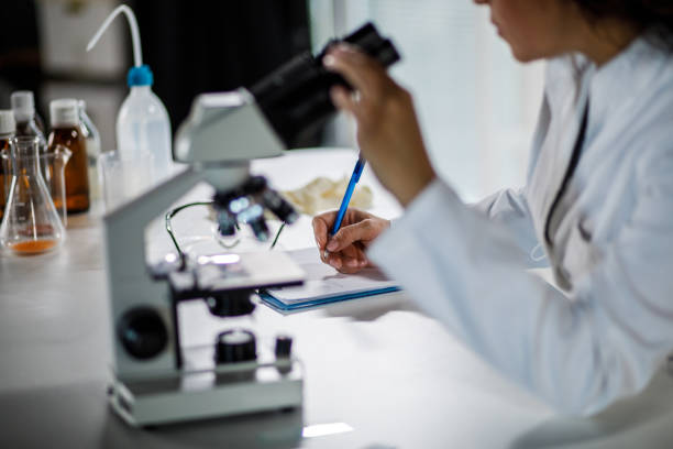 Cut out shot of female scientist writing notes on a clipboard after analyzing samples under a microscope stock photo