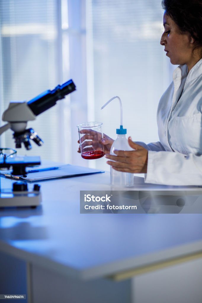 Female scientist adding liquid into a beaker with wash bottle she is using Candid shot of mid adutl female scientist using a wash bottle and adding liquid into a beaker with red powder in it while doing experiments at the laboratory. 35-39 Years Stock Photo