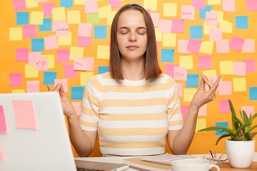 Indoor shot of relaxed beautiful Caucasian woman wearing striped T-shirt posing against yellow wall with colorful memo cards, practicing yoga on workplace, sitting in front laptop.