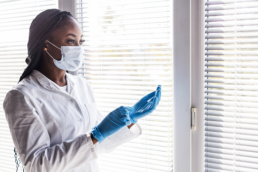 Copy space shot of serious young female doctor standing by the window, taking off protective gloves and looking outside.