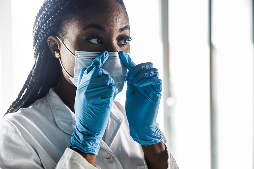 Portrait of serious young female laboratory technician adjusting the protective face mask to better fit her face before working on a project.