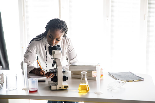 Portrait of diligent young female student working at the laboratory, writing notes and studying after analyzing samples under microscope.