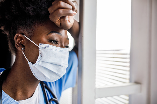 Copy space shot  of overworked female doctor, wearing a protective face mask, leaning against the window in her office, looking outside, taking a break from seeing all the patients.